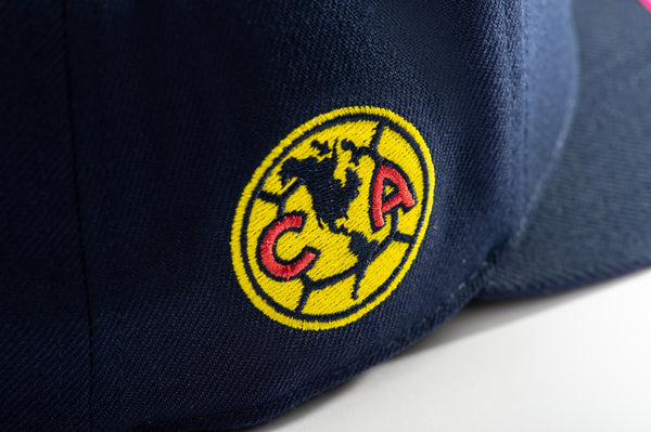 Club America Day of the Dead Snapback Hat with America script with marigold floral overlay embroidered on the front, Club America logo embroidered on the side, "Dia de Muertos" embroidered on the back, and marigold floral print on the under peak. The hat has a high crown, flat peak brim, and a snapback closure.